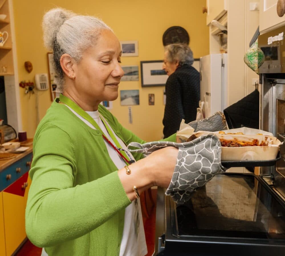 An older female adult with grey hair and wearing green putting pasta in the oven inside the litchen