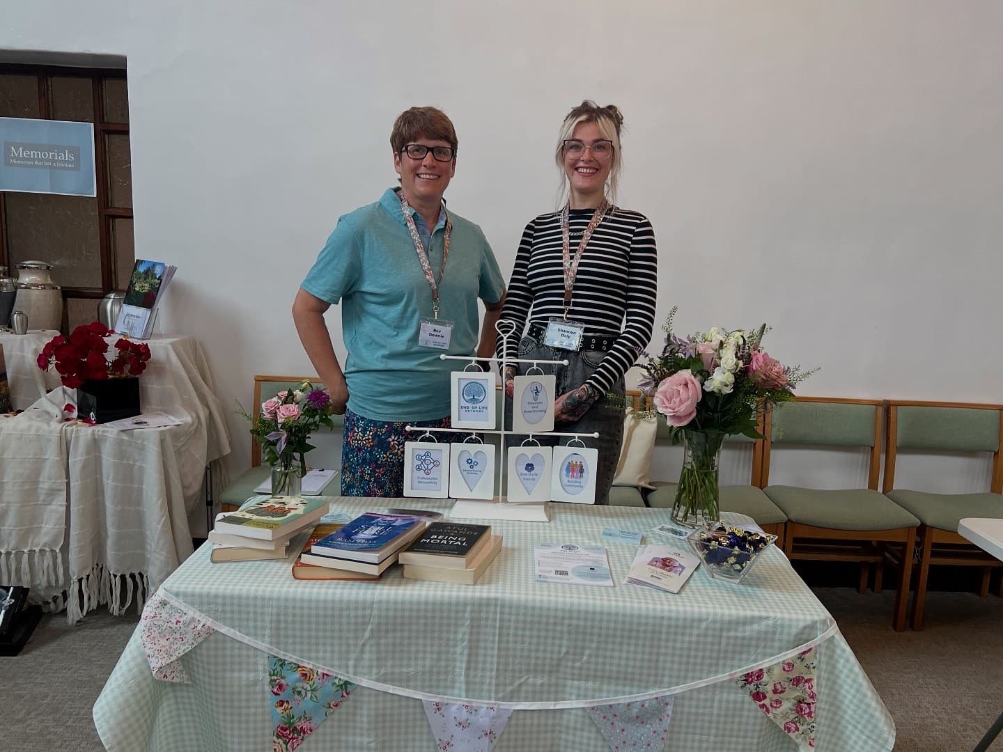 Two smiling people stand behind a table with books, flowers, and cards at an indoor event. - Home Instead