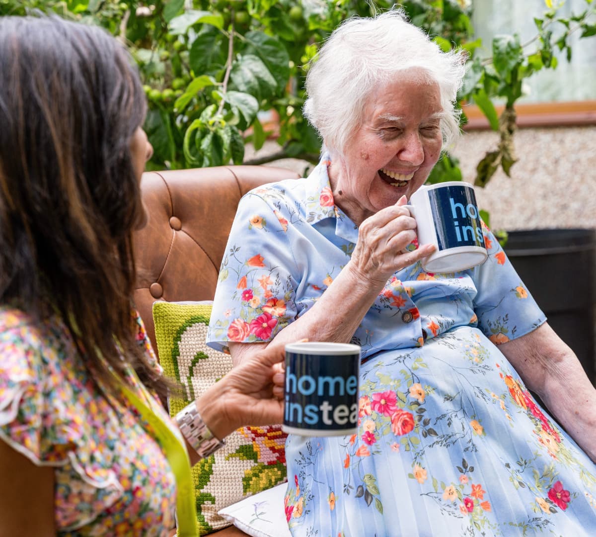 An older female adult with white hair happy and laughing while drinking coffee in the garden