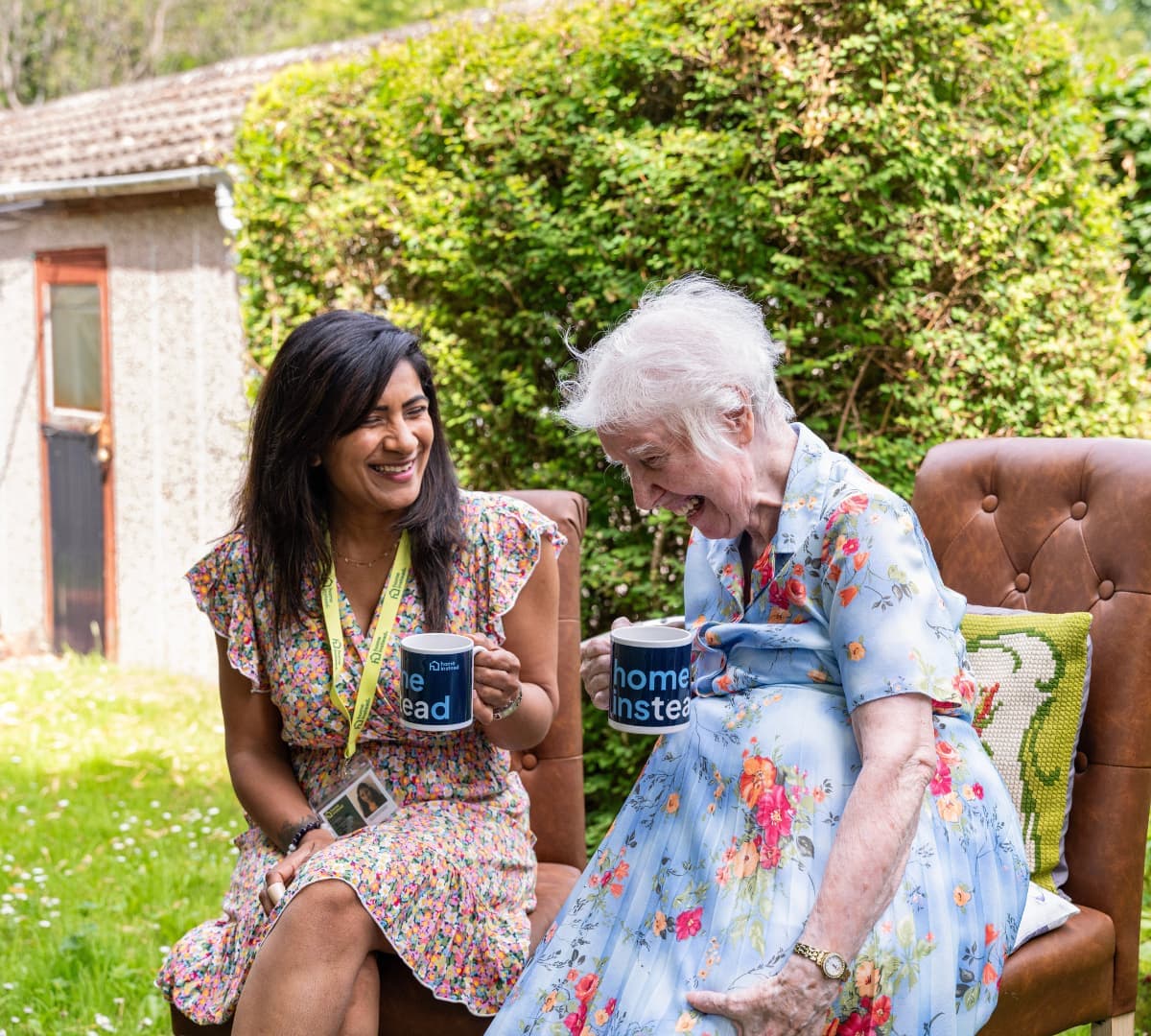 Two women having coffee while sitting on a chair in the garden, both happy and laughing