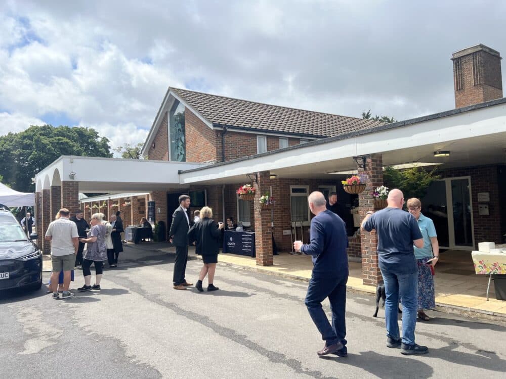 People gather and chat outside a brick building on a cloudy day, with hanging baskets and tables visible. - Home Instead