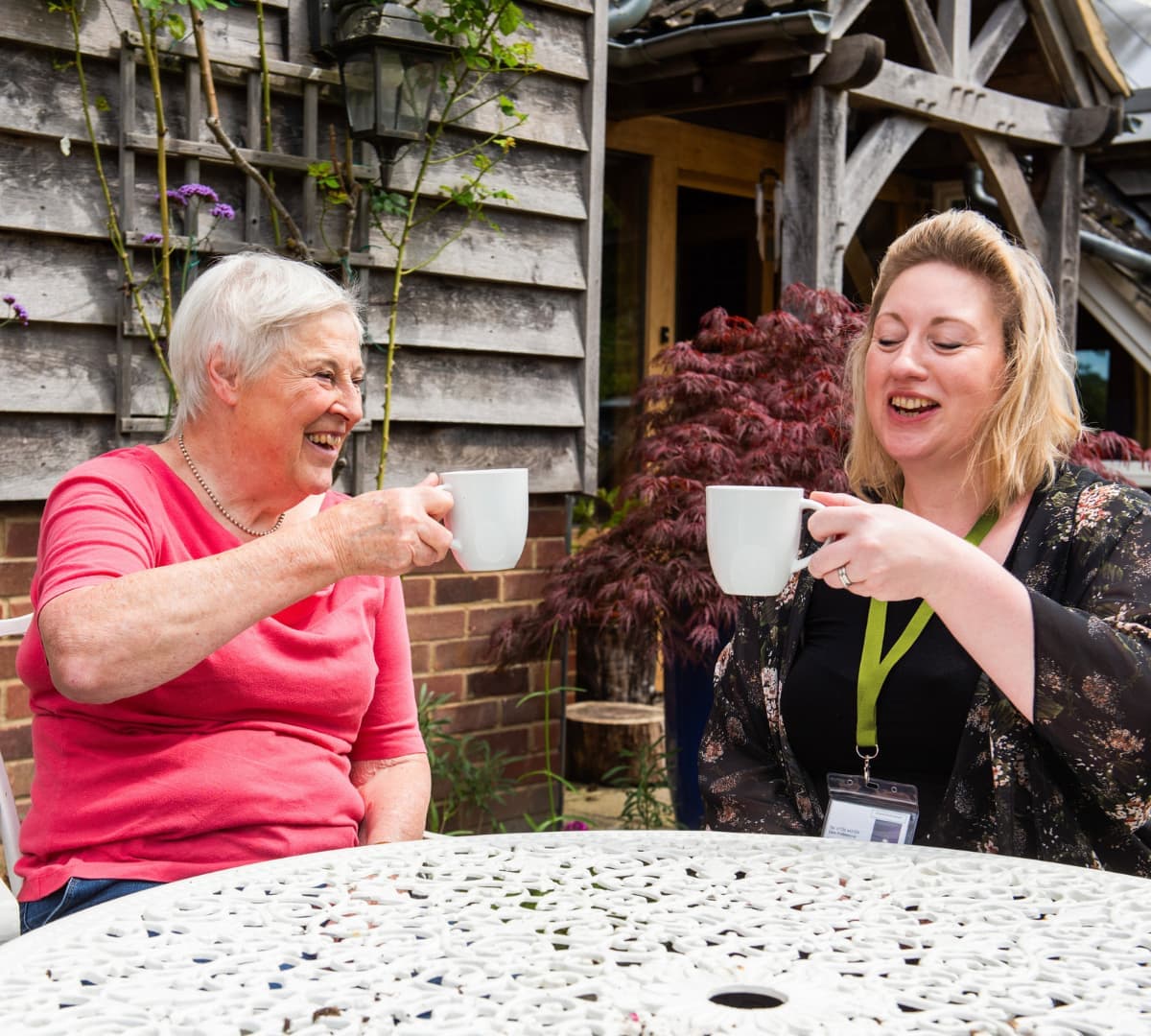 Two women toasting using a cup of coffee in the garden both happy and smiing