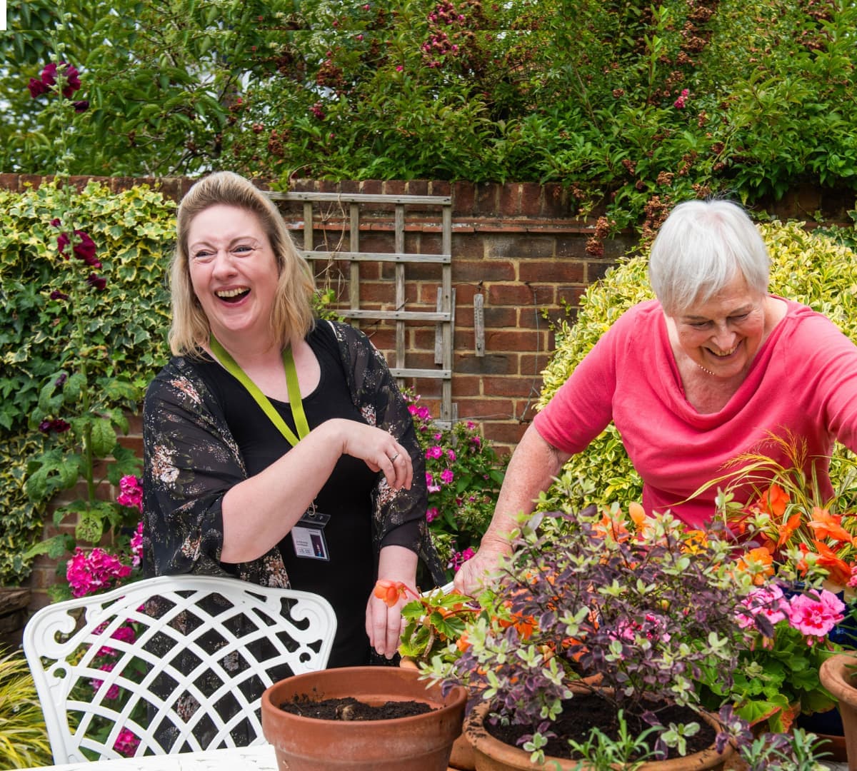 Two women laugh together while gardening, surrounded by colorful flowers and greenery outdoors. - Home Instead