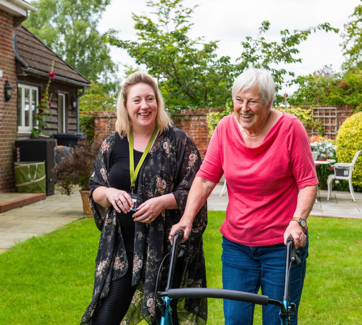 An older female adult with grey hair and wearing pink walking in the garden using a walker with her younger carer with blonde hair and wearing long dress both happy and smiling and walking inside the garden with green grass
