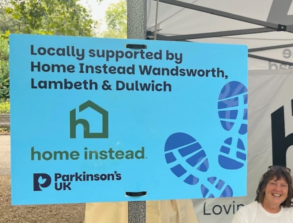 Blue sign reads "Locally supported by Home Instead Wandsworth, Lambeth & Dulwich" with logos; woman smiles nearby. - Home Instead