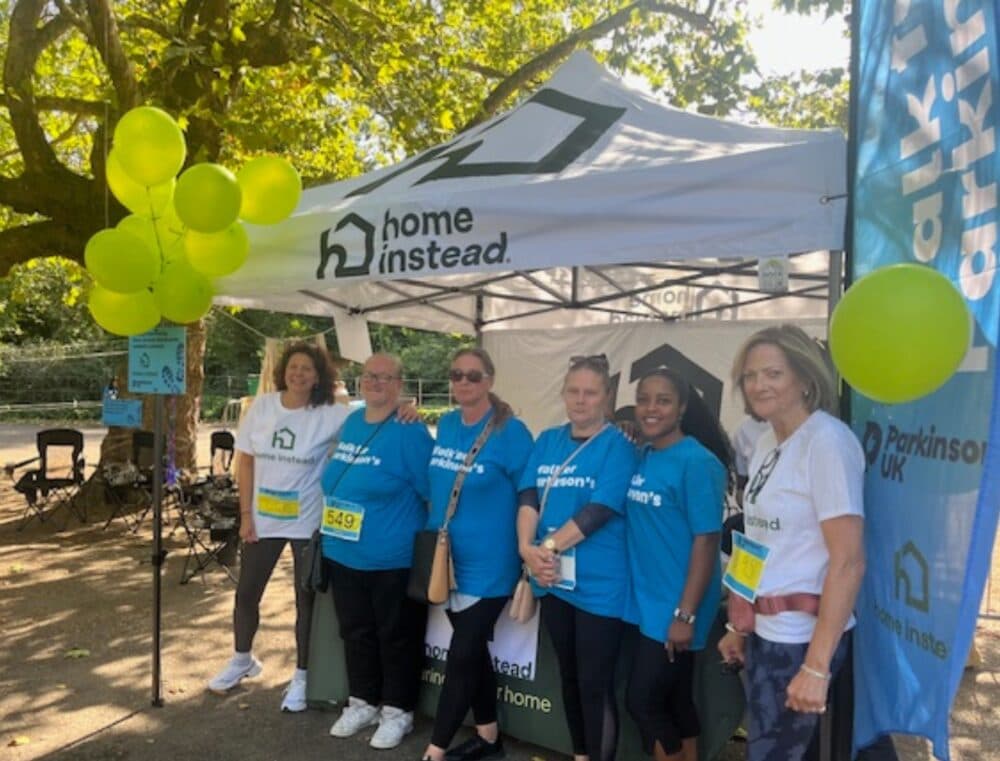 Six women stand in front of a Home Instead tent at an outdoor event, wearing event t-shirts and smiling. - Home Instead