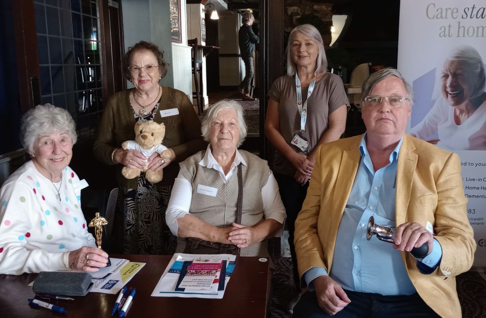 Five older adults, three seated and two standing, pose indoors holding small awards and a teddy bear. - Home Instead