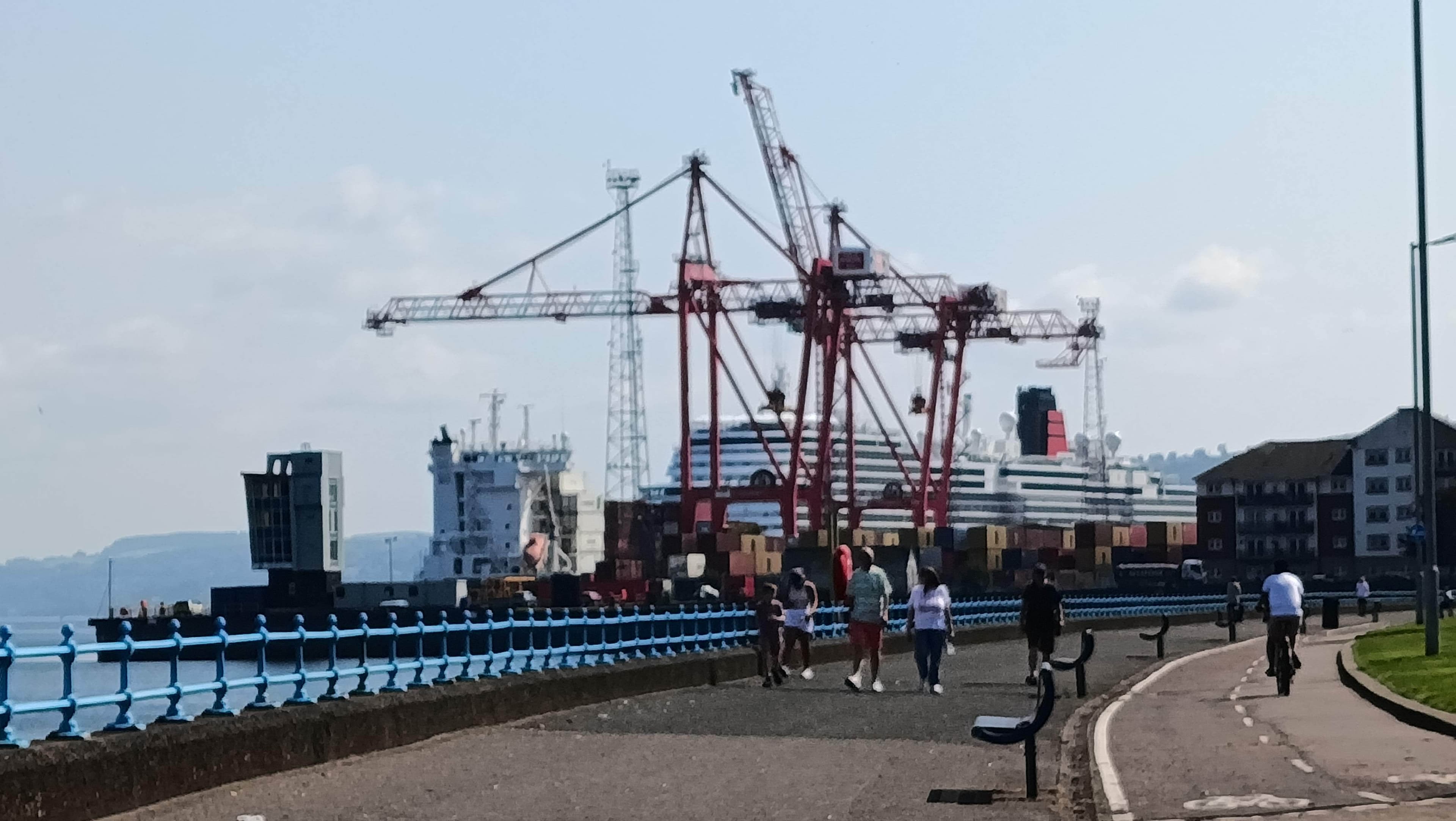 People walk and cycle along a waterfront path near cranes, shipping containers, and a docked ship. - Home Instead