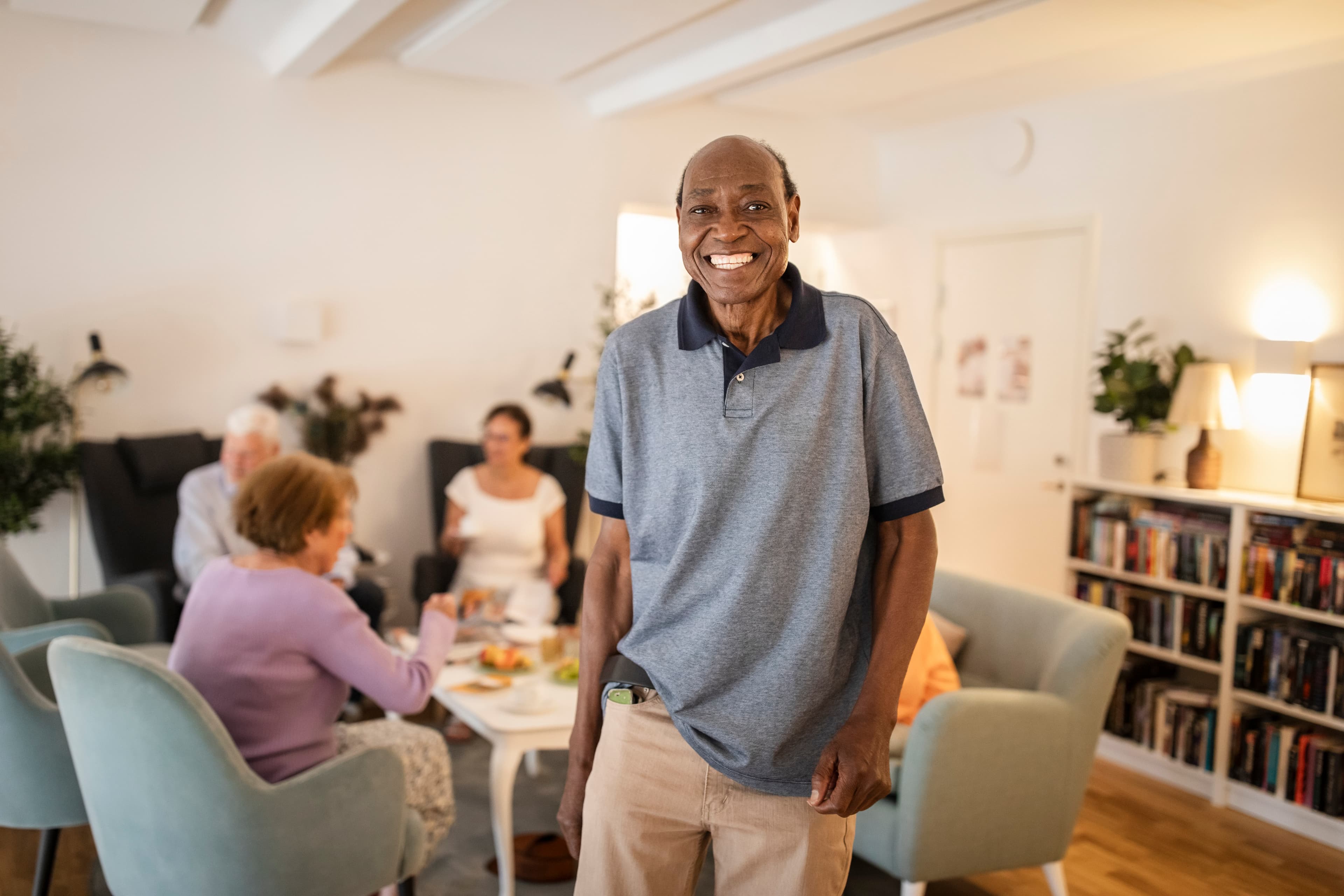 Smiling man stands in a living room with people sitting and talking at a table in the background. - Home Instead