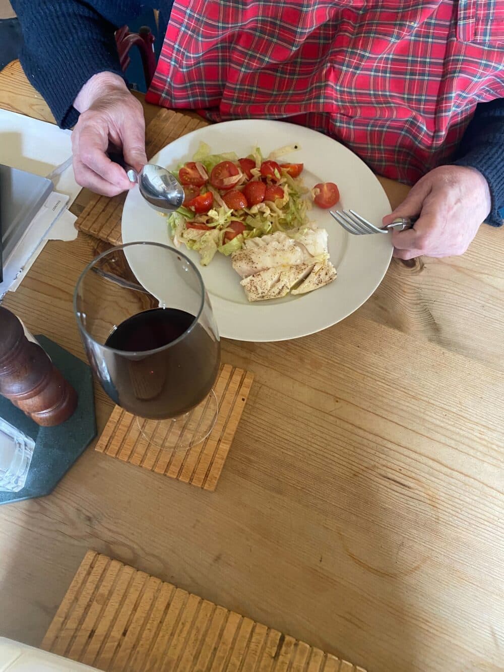 Person in plaid apron eating salad and chicken with a glass of red wine on a wooden table. - Home Instead