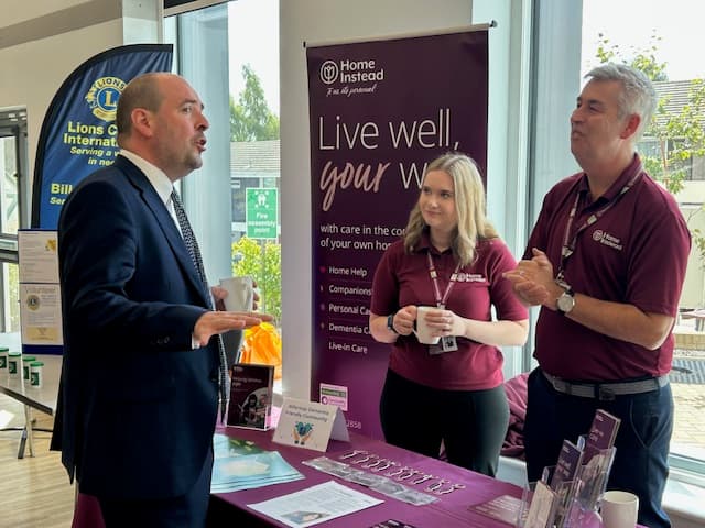 Three people talk at a care service booth with “Live well, your way” displayed on a banner behind them. - Home Instead