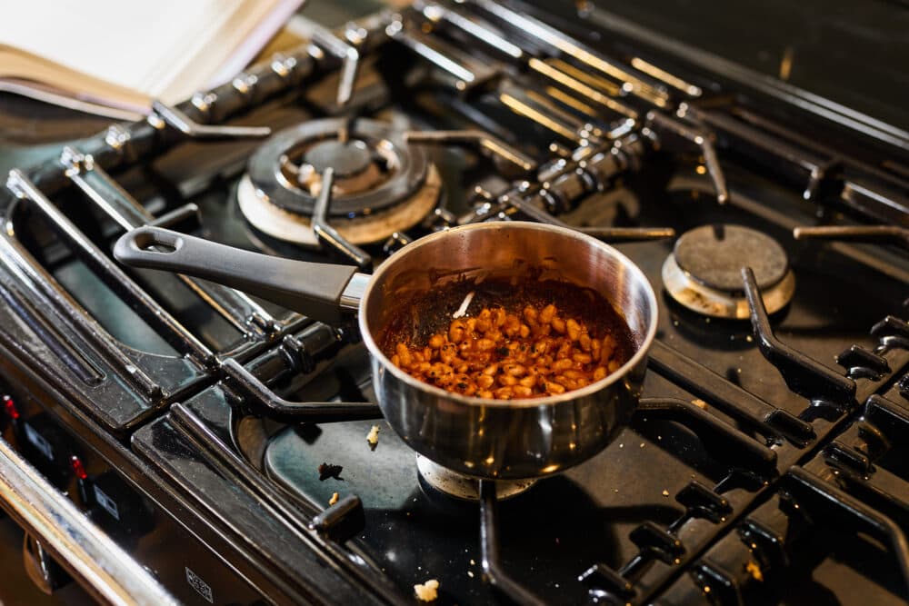 A saucepan of baked beans simmers on a gas stove with some crumbs nearby. - Home Instead