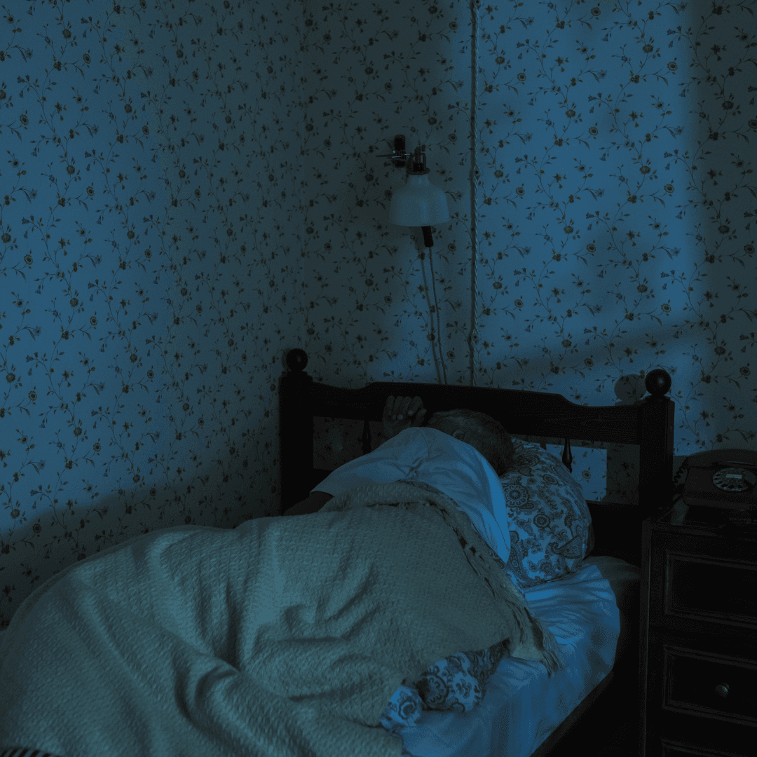 A person lies in bed under a blanket in a dimly lit room with floral wallpaper. - Home Instead