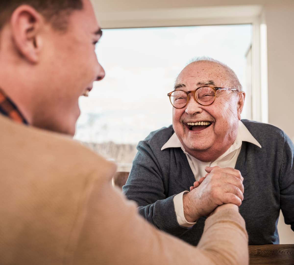 Happy older adult with grey hair and wearing eyeglasses laughing with his carer inside the house