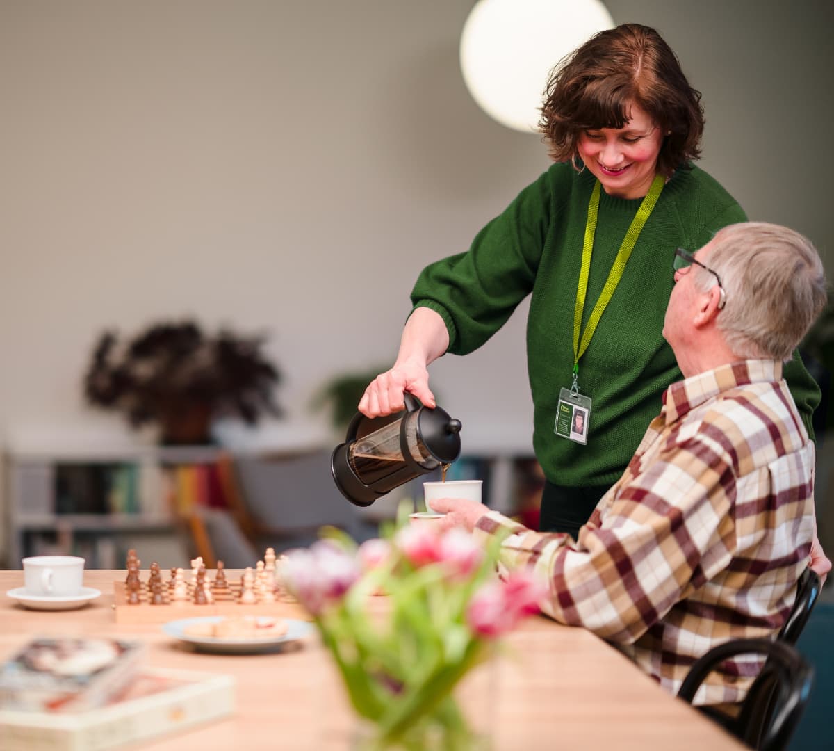 A woman pours coffee for an older man seated at a table with a chessboard and flowers. - Home Instead