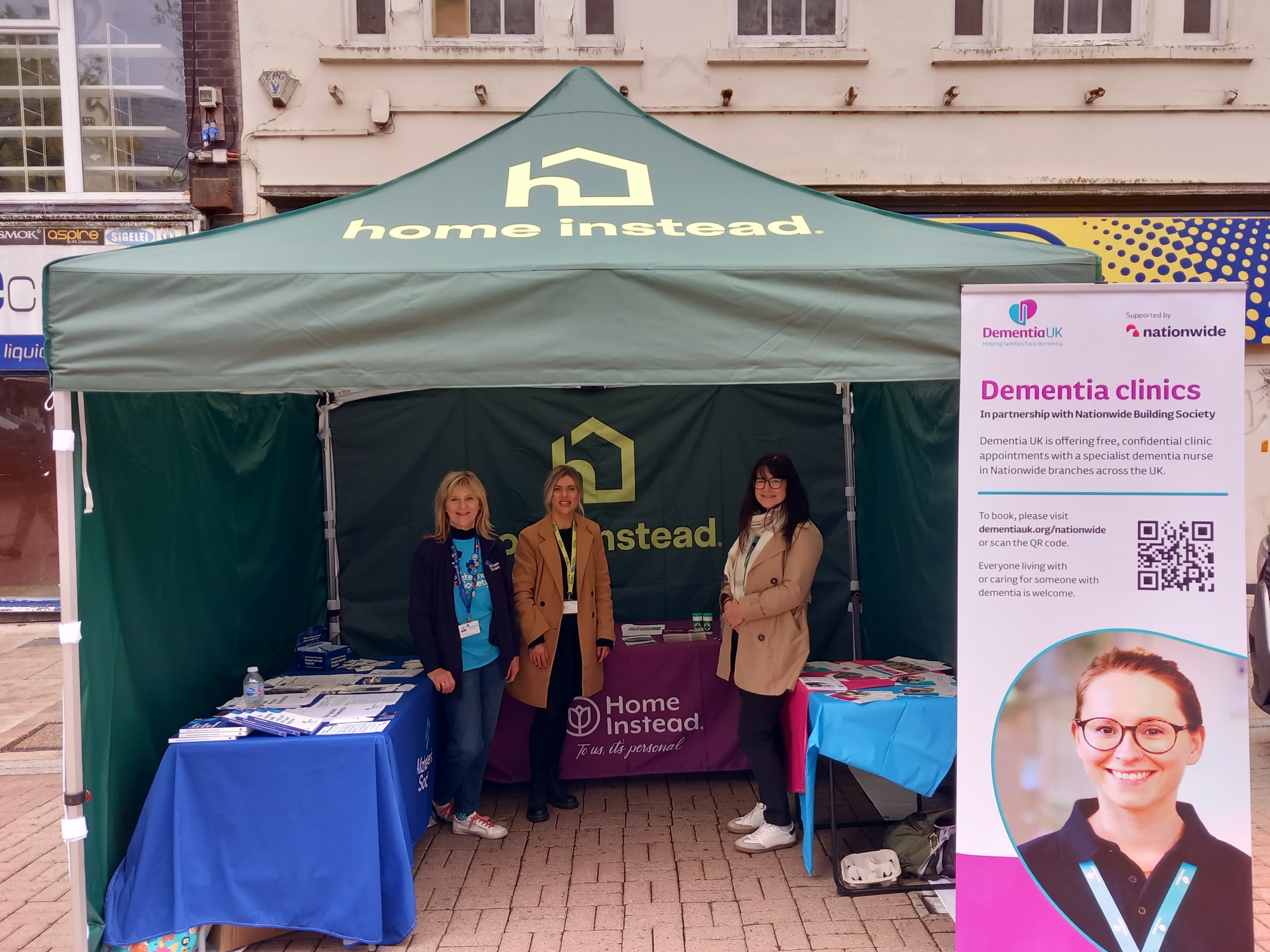Three women stand by a green Home Instead tent and Dementia clinics sign at an outdoor event. - Home Instead