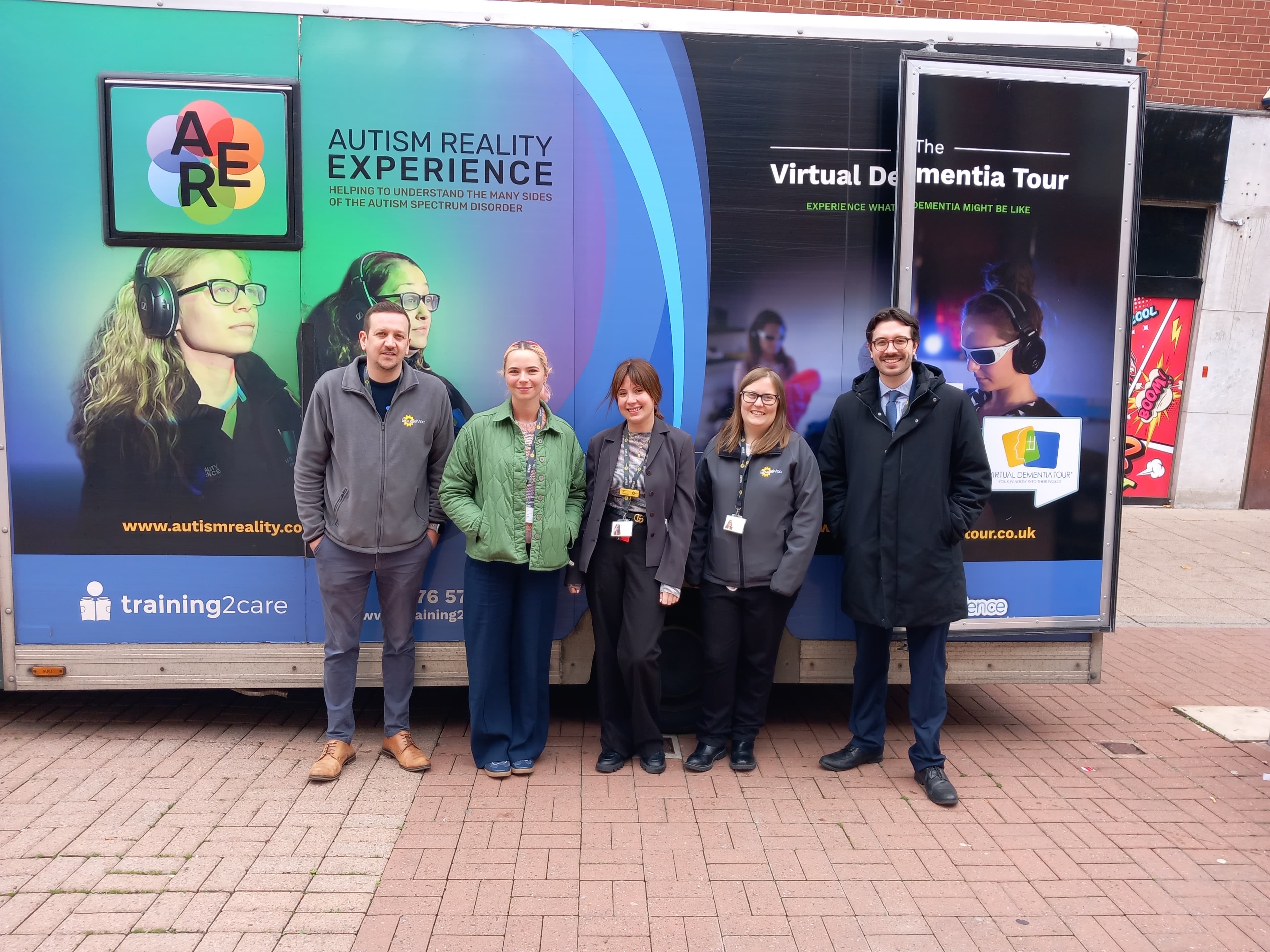 Five people standing and smiling in front of an Autism Reality Experience and Virtual Dementia Tour van. - Home Instead