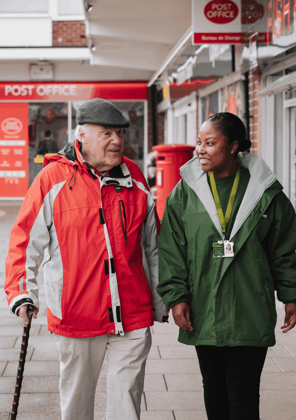 Community support worker walking with an older man outside a post office, both smiling and engaged in conversation.
