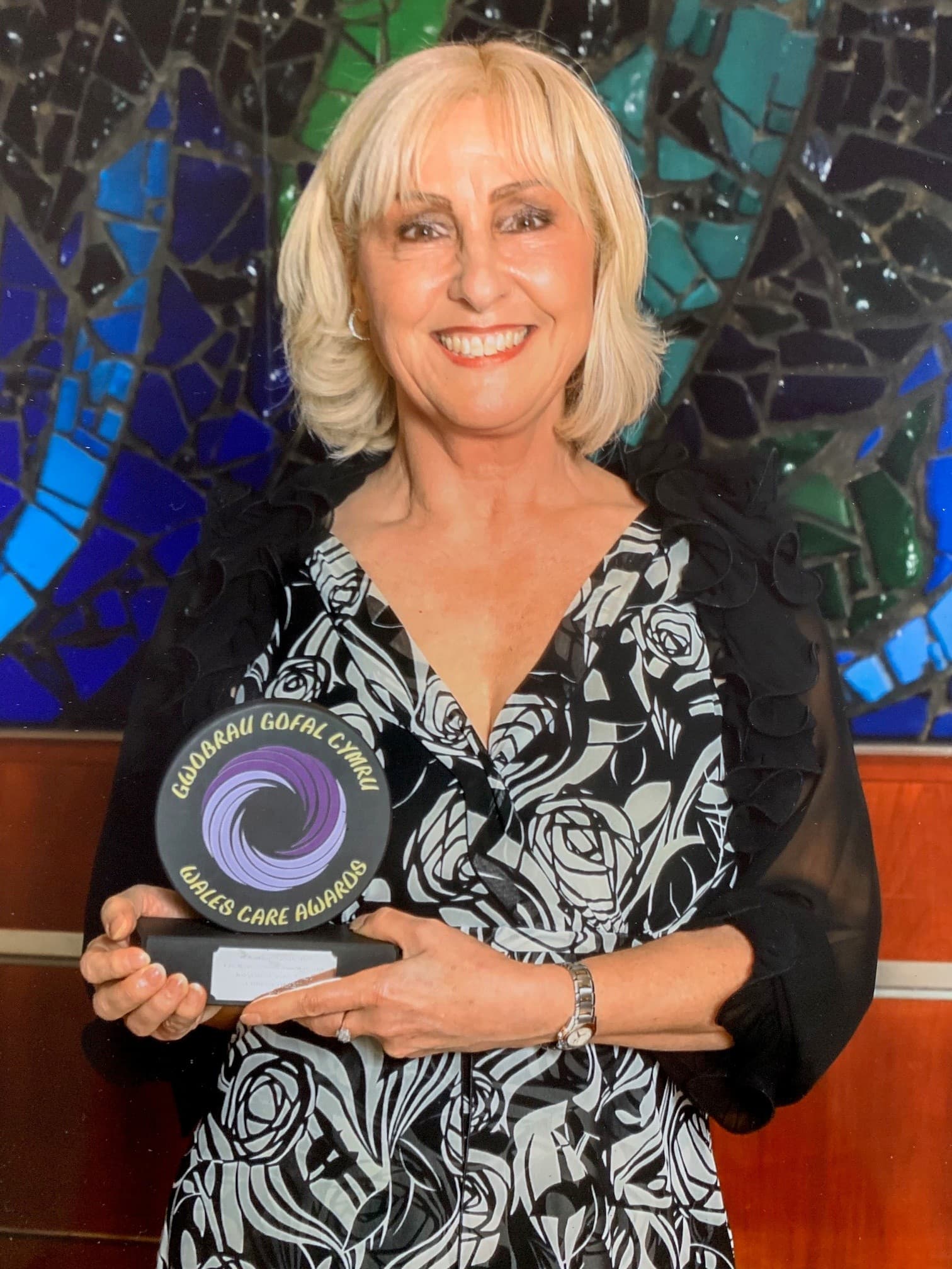 Smiling woman holding a "Carers Care Awards" trophy in front of a colorful mosaic background. - Home Instead