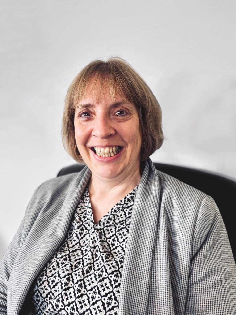 Smiling woman with short light brown hair wearing a patterned top and gray blazer, seated indoors. - Home Instead