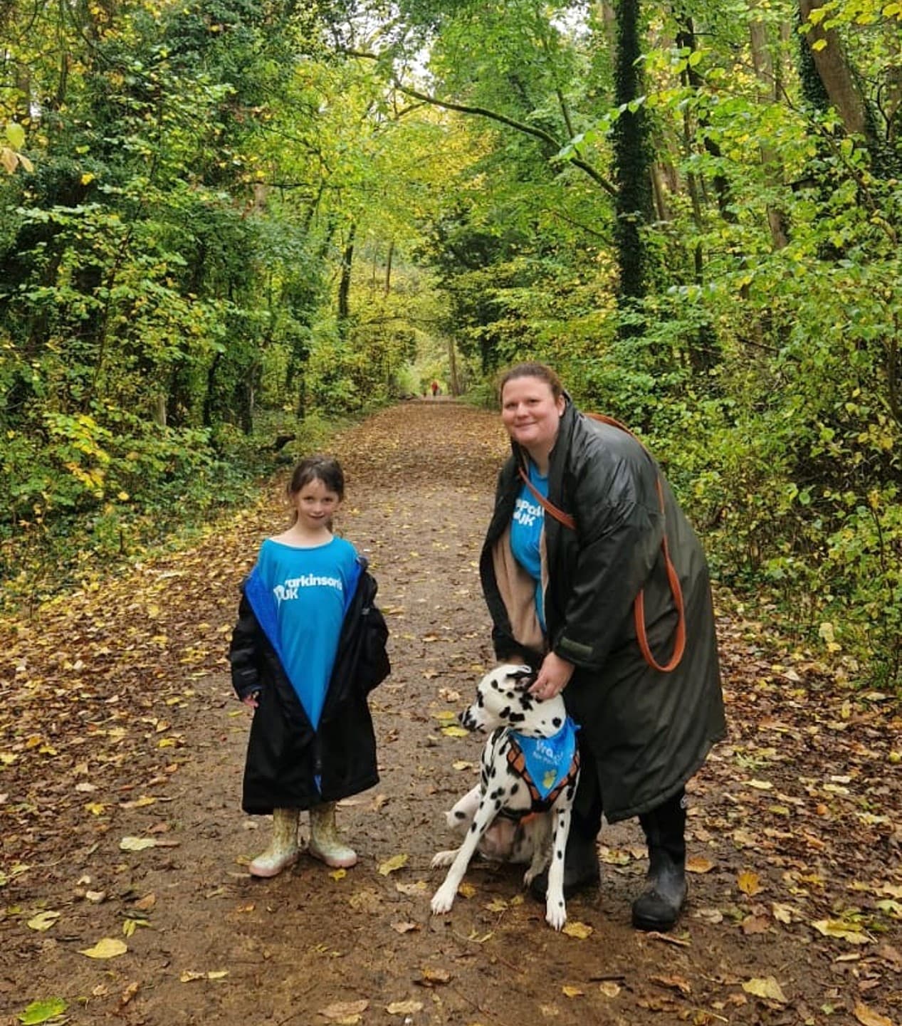 A woman and girl stand with a Dalmatian dog on a leafy forest path, all wearing blue shirts. - Home Instead
