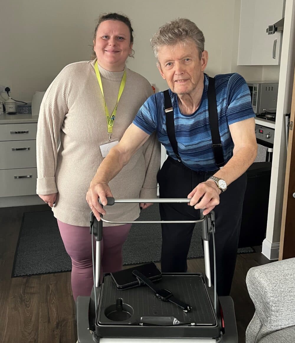 A smiling woman stands next to an older man using a walker in a kitchen setting. - Home Instead