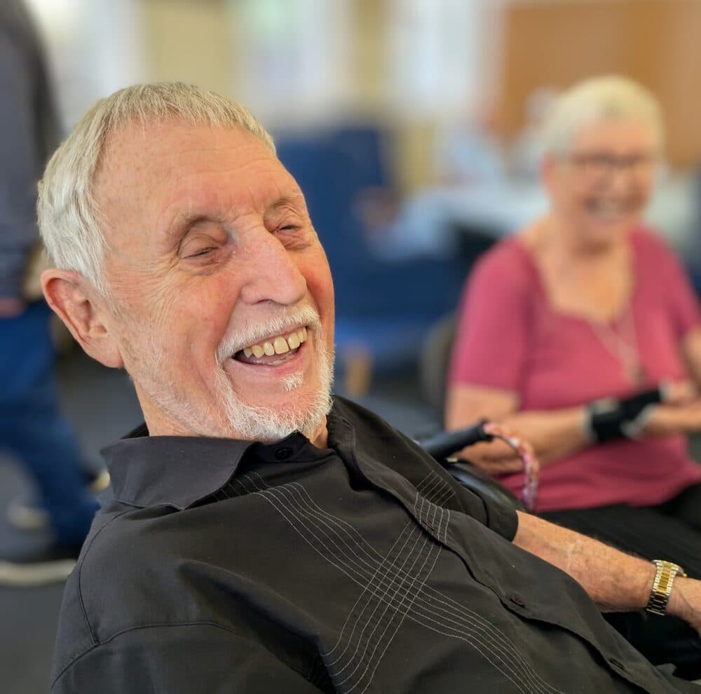 An elderly man in a black shirt smiles while sitting next to an elderly woman in a pink shirt. - Home Instead