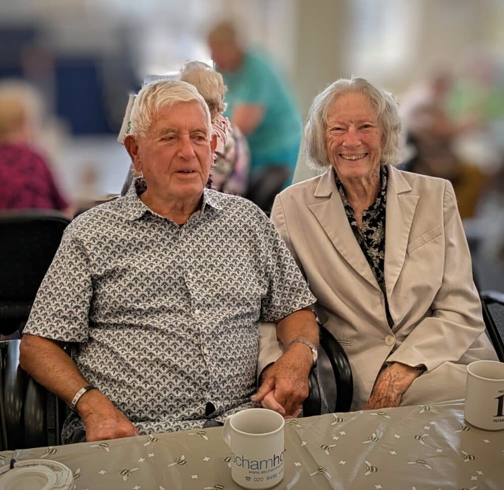Elderly man and woman smiling while sitting together at a table with mugs in front of them. - Home Instead