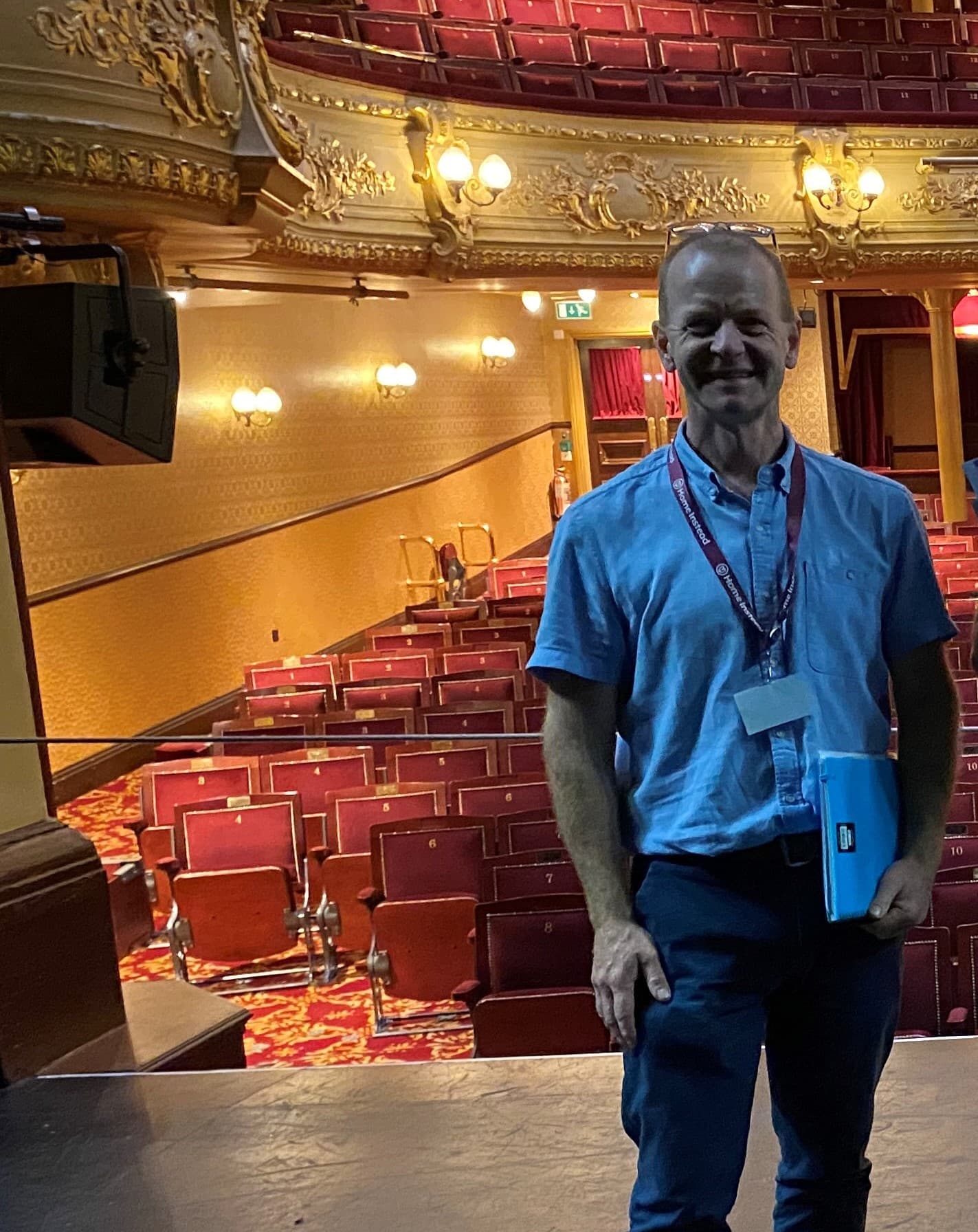 Man standing on stage in an ornate, empty theater with red seats and golden decor, holding a blue folder. - Home Instead