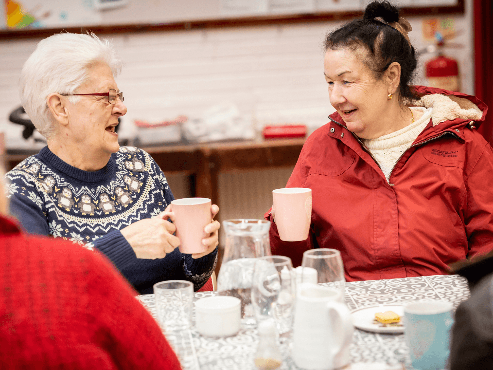 Two older women smile and chat while holding mugs at a table set with cups, glasses, and snacks. - Home Instead