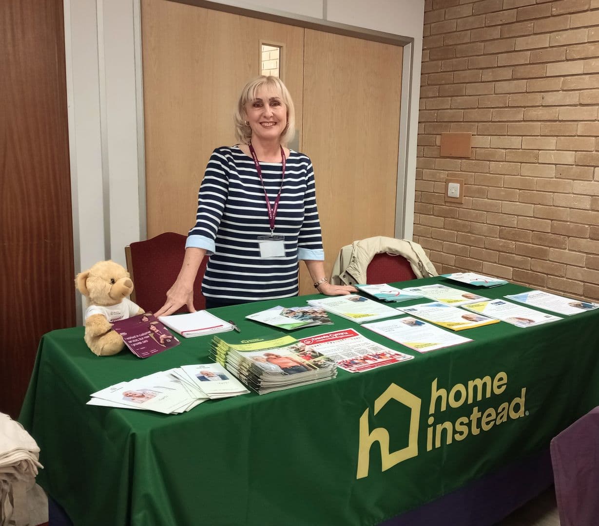 A woman stands behind a Home Instead table covered with brochures and a teddy bear, smiling at the camera. - Home Instead