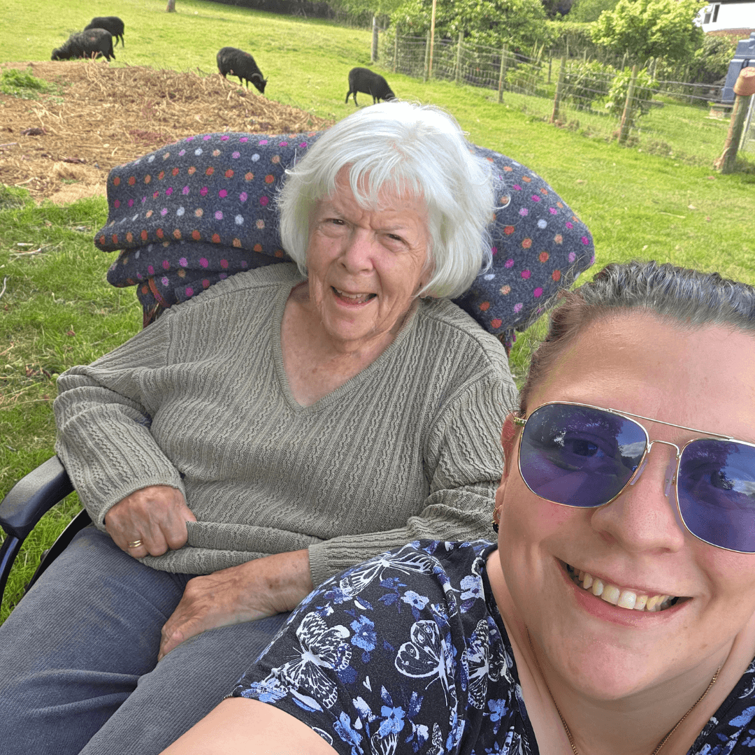 Older woman and younger woman smiling outside, with sheep grazing in the grassy background. - Home Instead