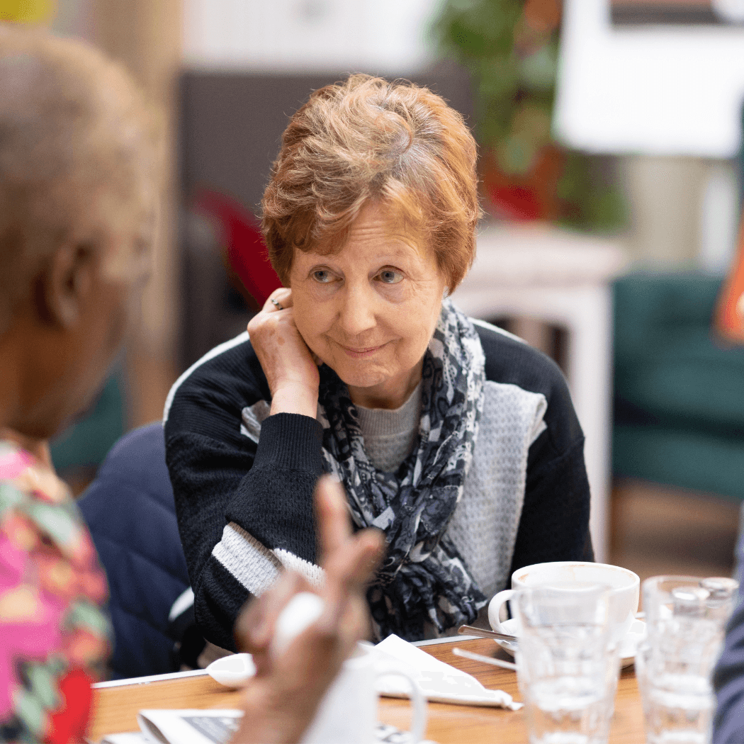 An older woman listens attentively to another person at a table with coffee cups and glasses. - Home Instead