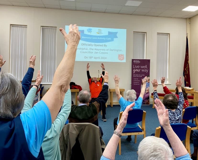 Seniors seated in a circle raise their arms during a group activity at a community center event. - Home Instead