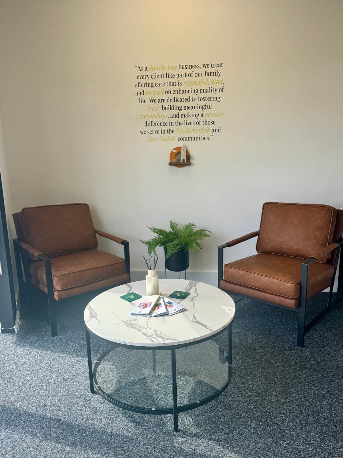 Two brown armchairs and a round white coffee table with magazines in a cozy office corner, with a quote on the wall. - Home Instead