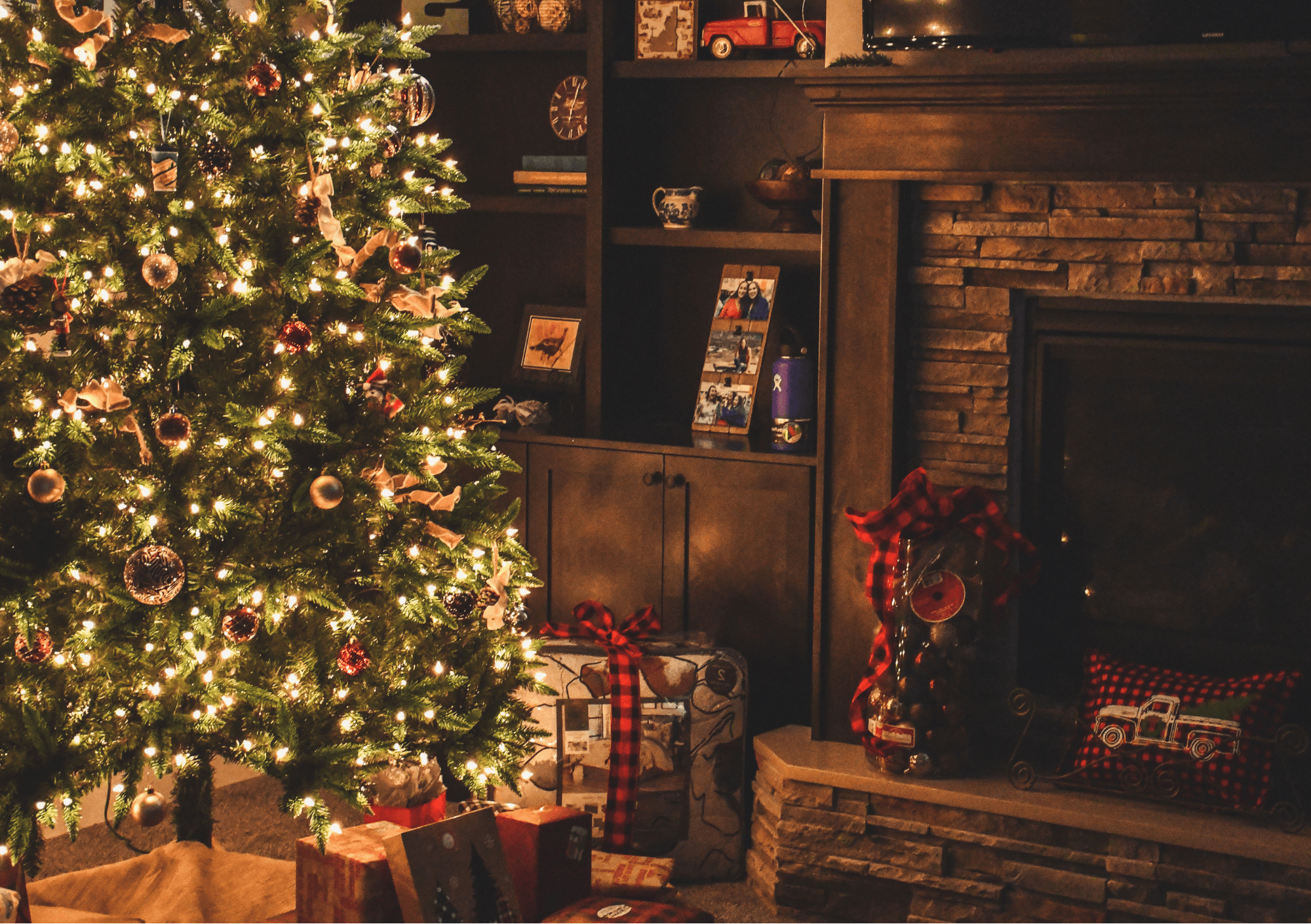 A decorated Christmas tree with lights and wrapped gifts beside a fireplace and shelves. - Home Instead
