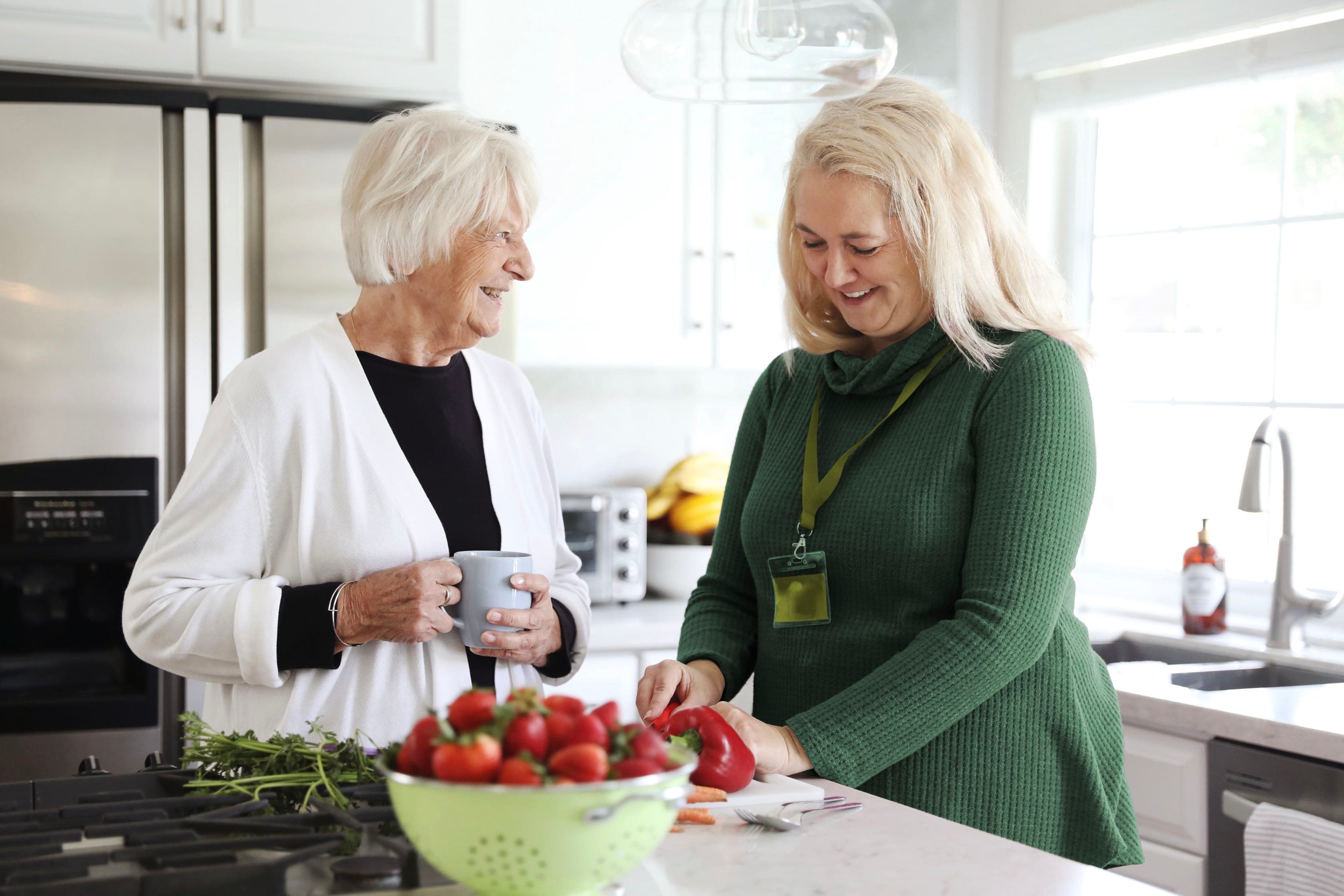 An elderly woman with a mug talks to a caregiver slicing vegetables in a bright kitchen. - Home Instead
