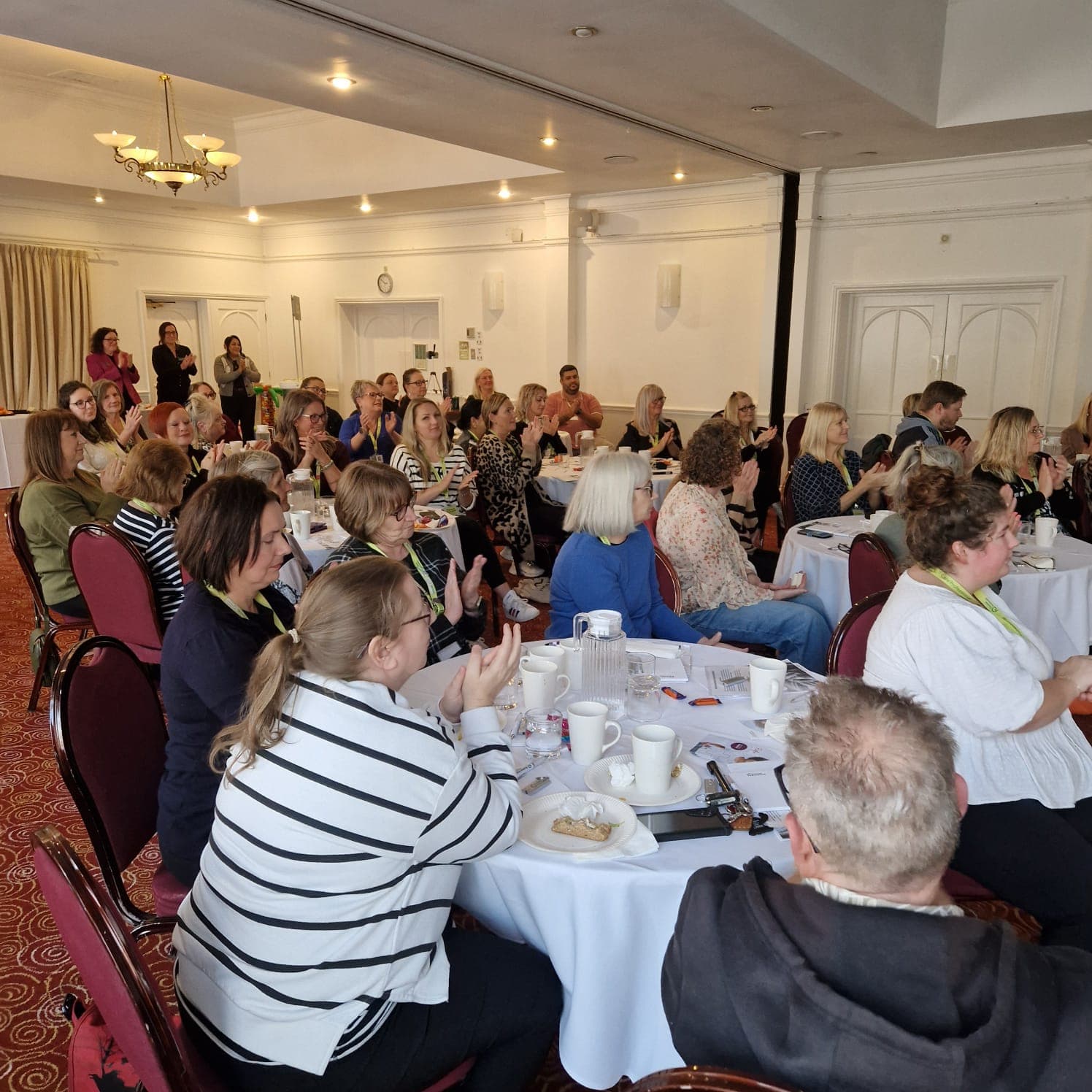 A group of people sit at round tables in a conference room, listening attentively to a speaker. - Home Instead