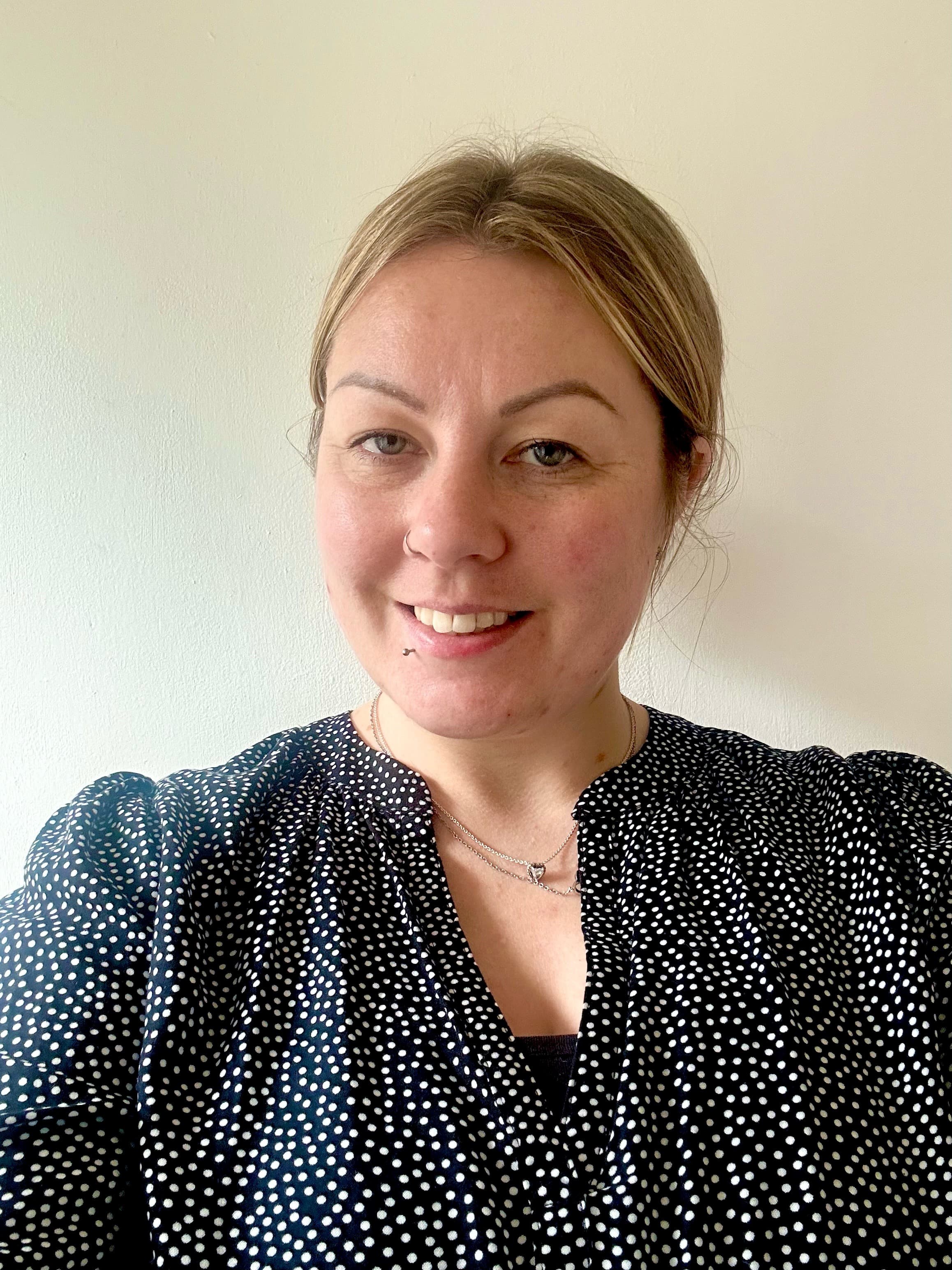 Smiling woman with blonde hair in a bun, wearing a black and white polka dot top, against a plain wall. - Home Instead