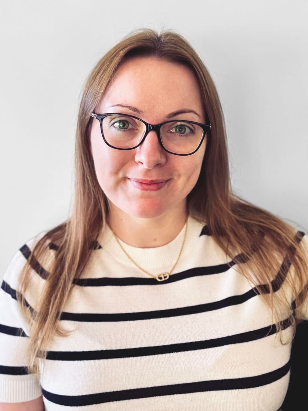 Woman with long blonde hair, glasses, and striped shirt smiling at the camera against a light background. - Home Instead