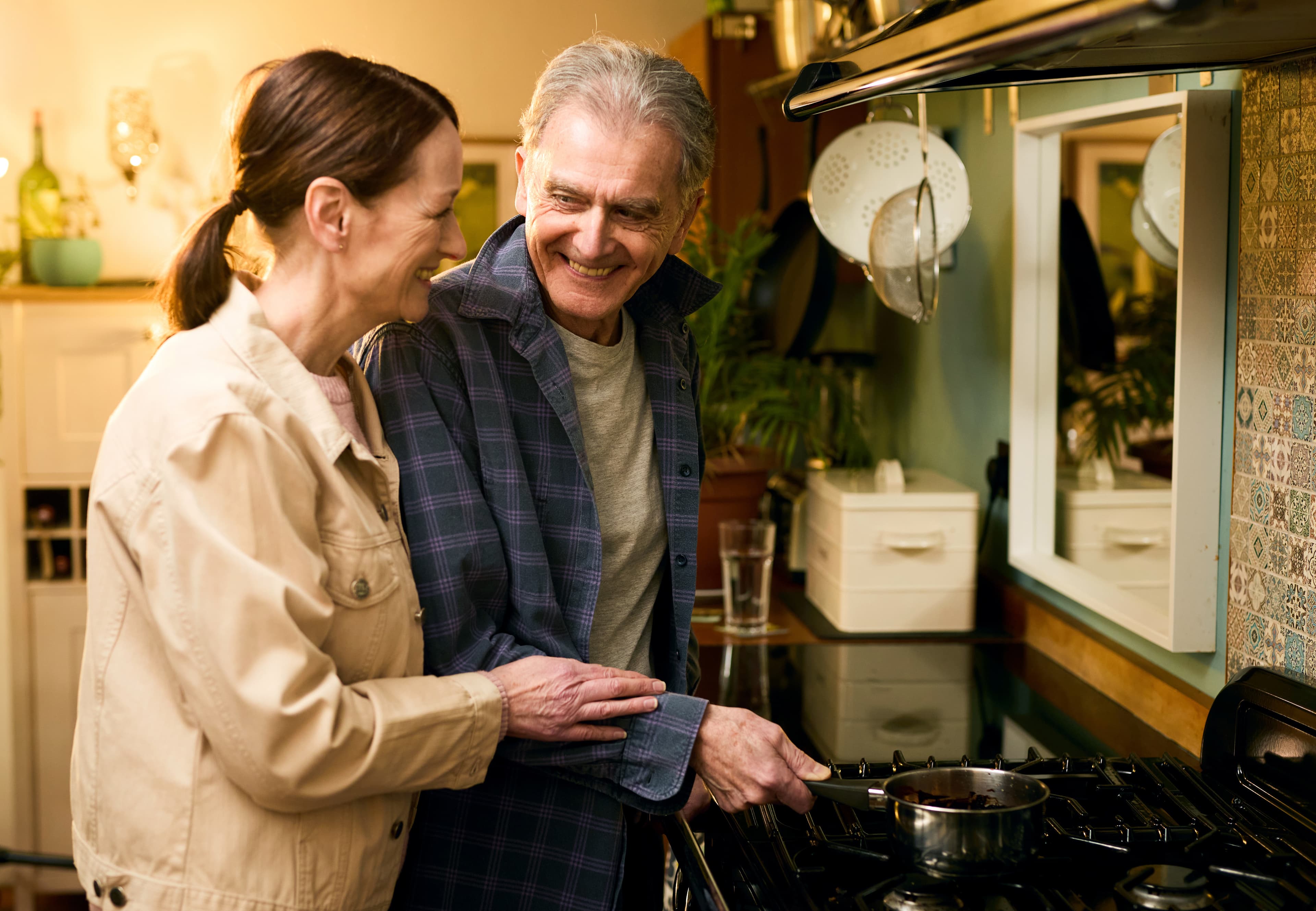 An older couple smiling and cooking together in a cozy kitchen. - Home Instead