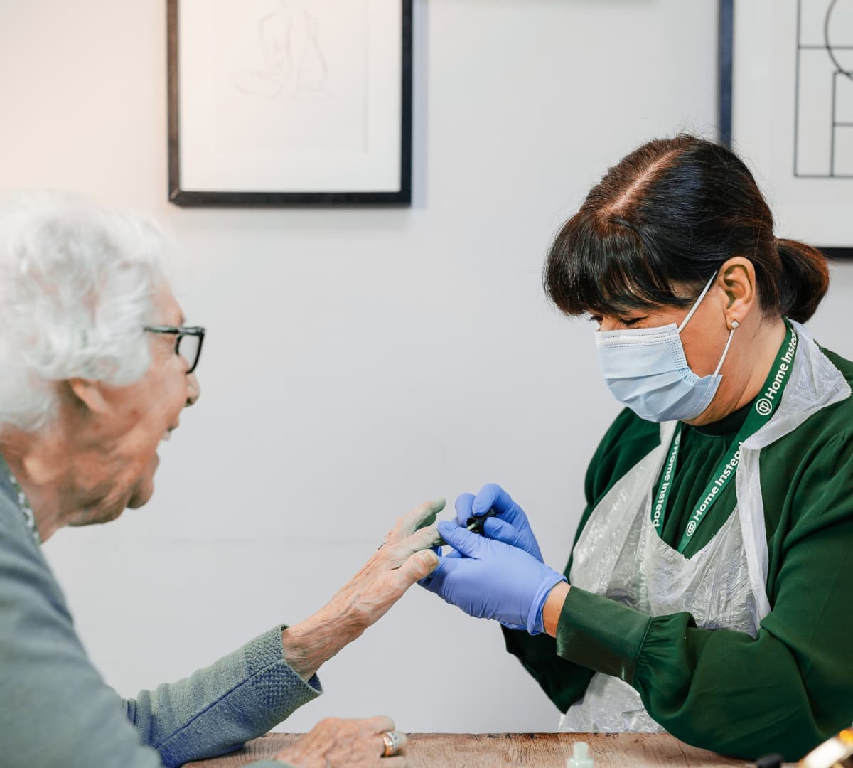 An older female adult with white hair and wearing eyeglasses having her nails done by a female carer wearing a mask and gloves and green dress with apron