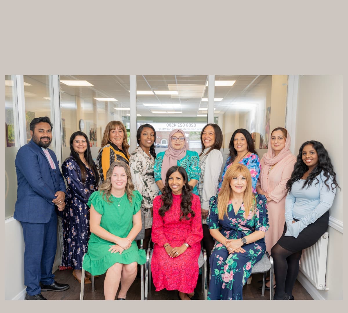 A group of thirteen adults, mostly women, pose and smile together in an office hallway. - Home Instead