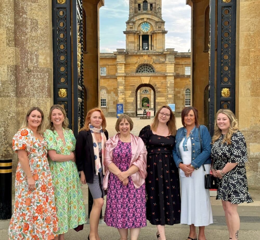 Seven women in colorful dresses pose and smile in front of a grand historic building with an arched gate. - Home Instead