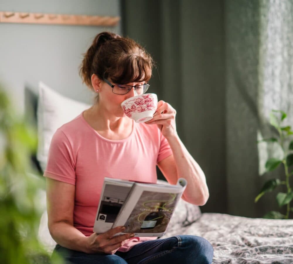 A woman sipping tea while reading and sitting on her bed inside the house
