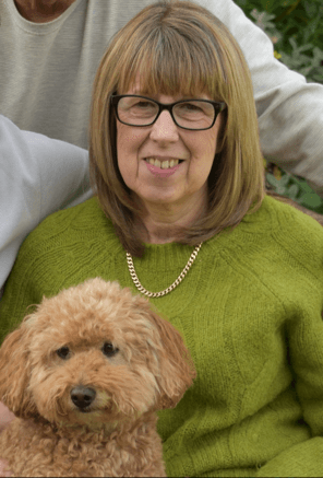 Woman in glasses and a green jumper smiling, holding a small curly-haired brown dog on her lap. - Home Instead