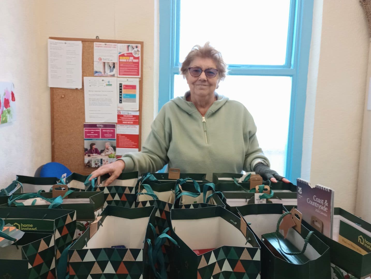 Smiling woman stands behind a table with several filled gift bags in a room with a bulletin board and window. - Home Instead