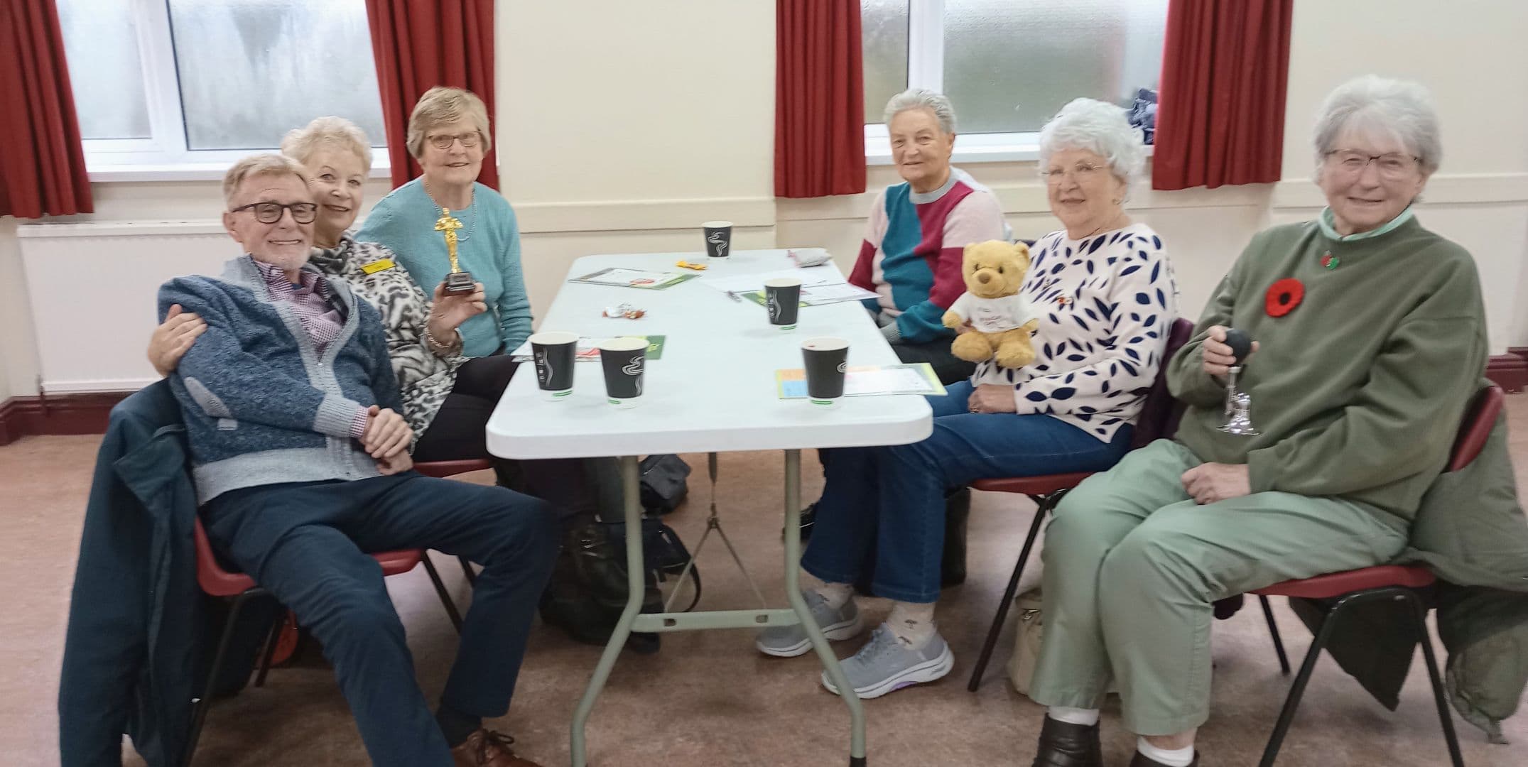 Seven older adults sit round a table with drinks, smiling; one holds a trophy, another a teddy bear. - Home Instead