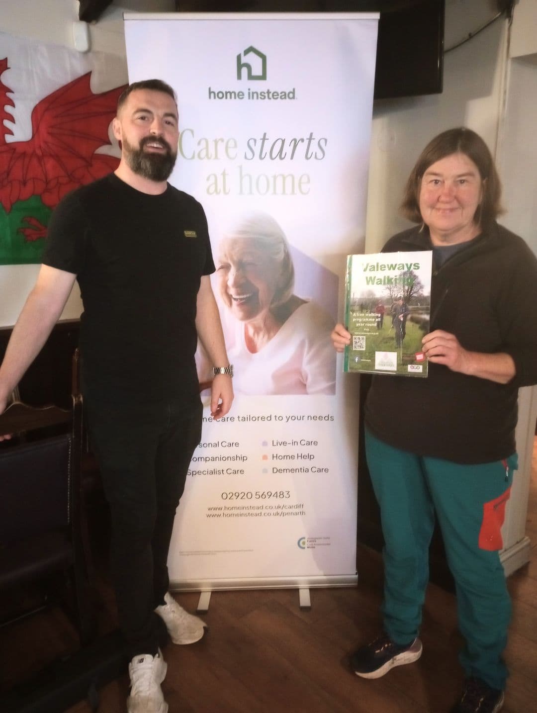 Two people stand by a "Care starts at home" banner; one holds a "Valeways Walks" booklet and smiles. - Home Instead
