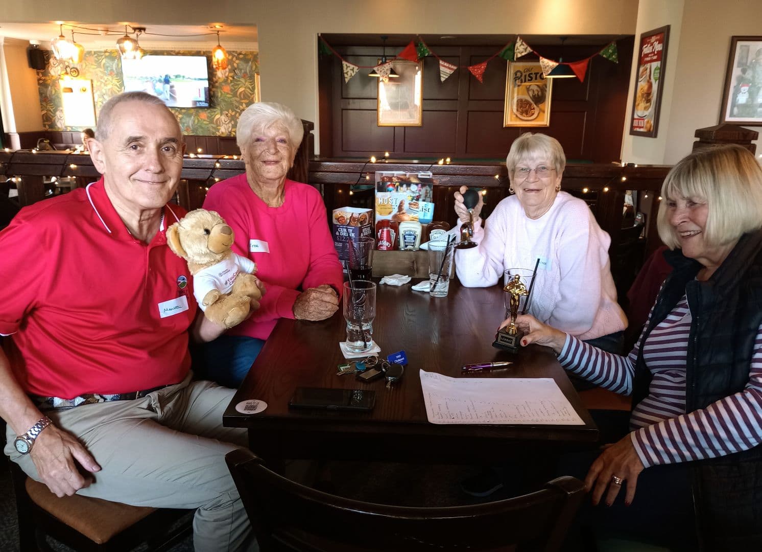 Four older adults sit around a pub table with drinks, papers, a trophy, and a teddy bear, smiling at the camera. - Home Instead